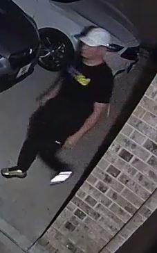 Camera footage photo of second suspect wearing black joggers, a black t-shirt with yellow print and a white ball cap.